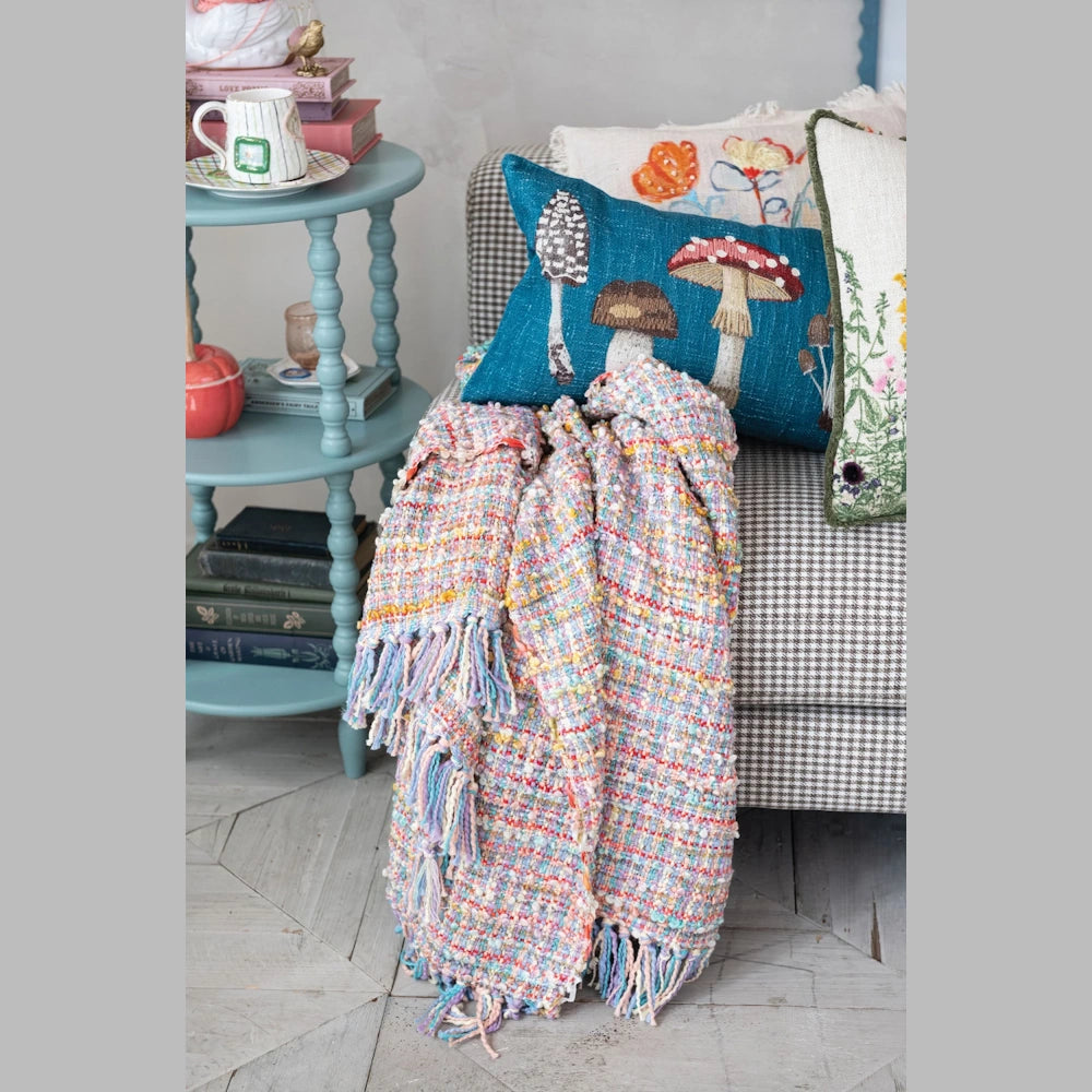 Colorful textured throw blanket with tassels draped on a chair near mushroom embroidered pillows and a blue side table.