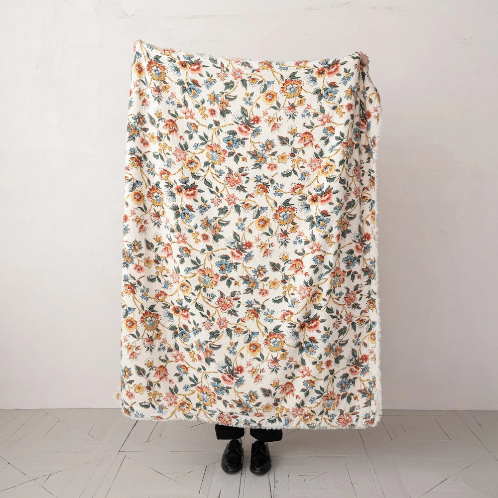 A large rectangular cream blanket featuring a dense floral vine pattern in orange, teal, and pink with frayed trim.
