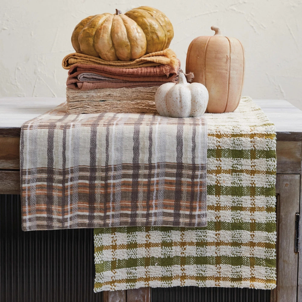 Plaid woven table runners in green and brown tones layered on a rustic wooden table with decorative pumpkins.