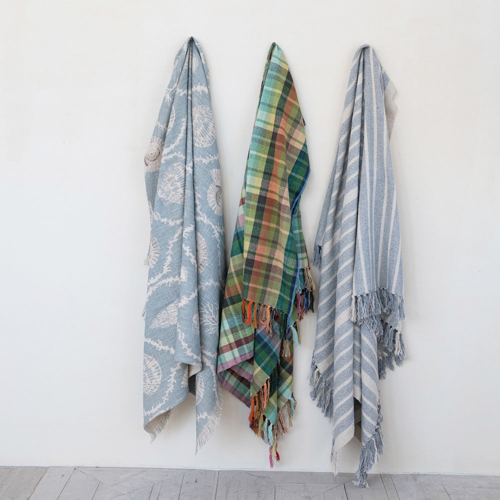 Three decorative woven cotton throw blankets with plaid, striped, and seashell patterns hanging on a wall.