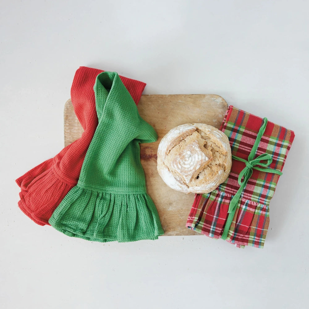Red, green, and plaid ruffled kitchen towels displayed on a wooden cutting board beside a loaf of bread.