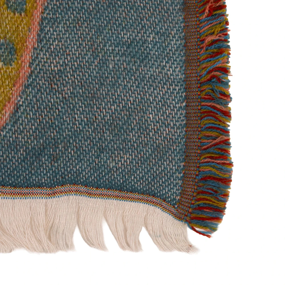 Close-up of a teal woven throw blanket showing intricate fabric texture, white fringe, and multicolored trim.