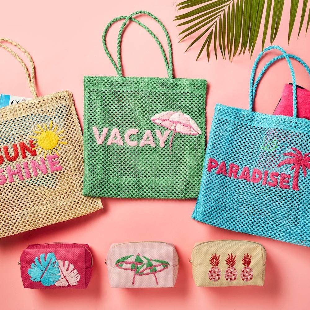 An assortment of woven summer tote bags and matching small pouches with tropical embroidery on a pink background.