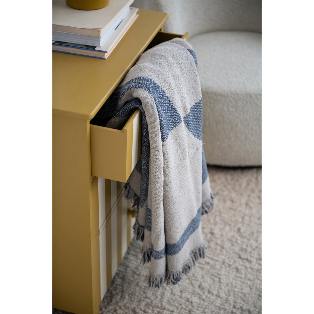 A fringed blue and grey patterned throw blanket draped over an open drawer of a mustard yellow nightstand.