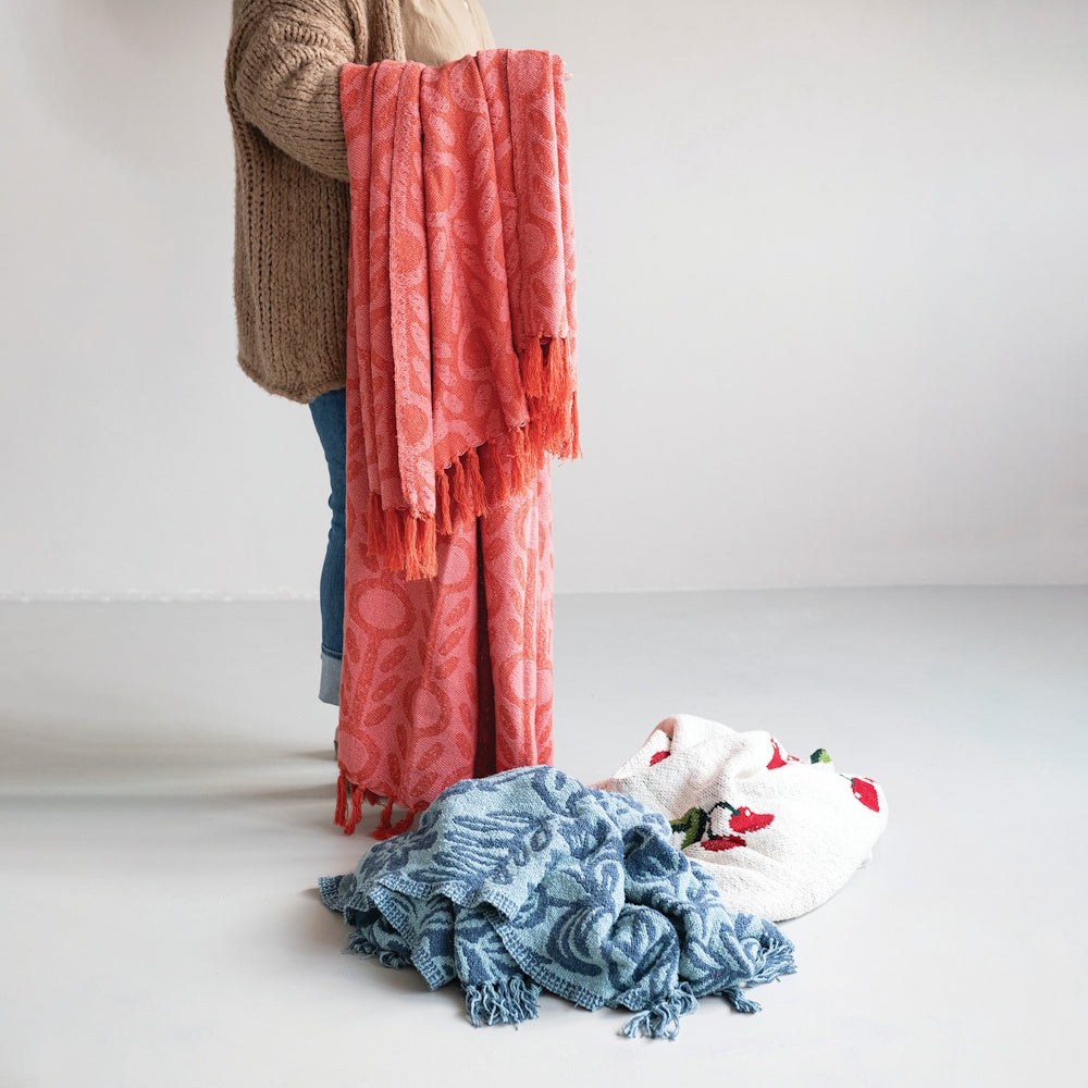 Woven recycled cotton blend jacquard throw in coral pink with a floral pattern and fringe, held up against other throws on the floor.