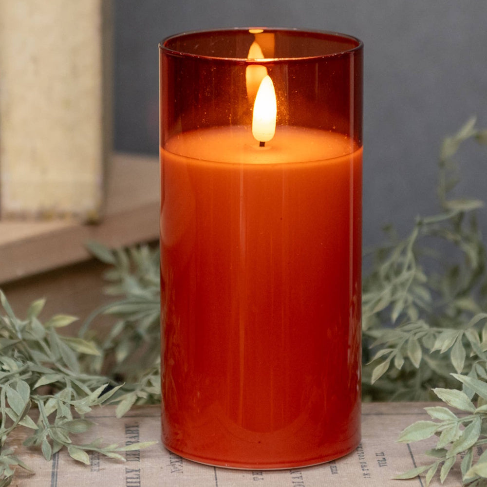 Large lit sienna-orange glass candle sitting on vintage paper surrounded by leafy greenery.