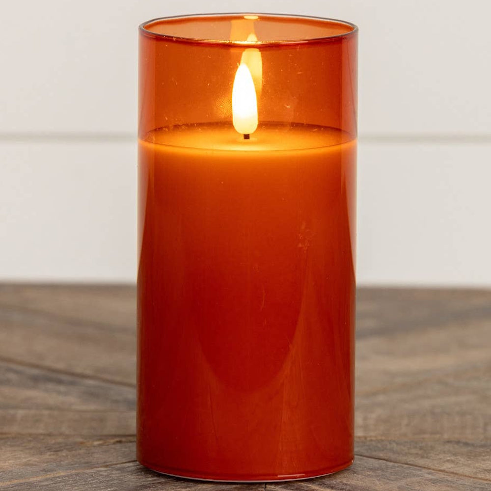 Tall sienna glass flameless candle glowing brightly on a rustic wooden surface.