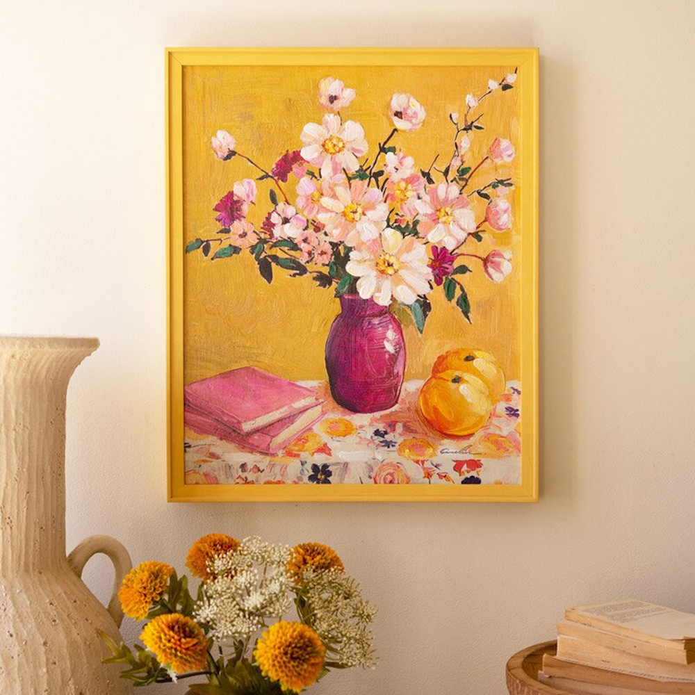 Still life painting in a yellow frame featuring pink flowers in a purple vase, books, and fruit on a yellow background.