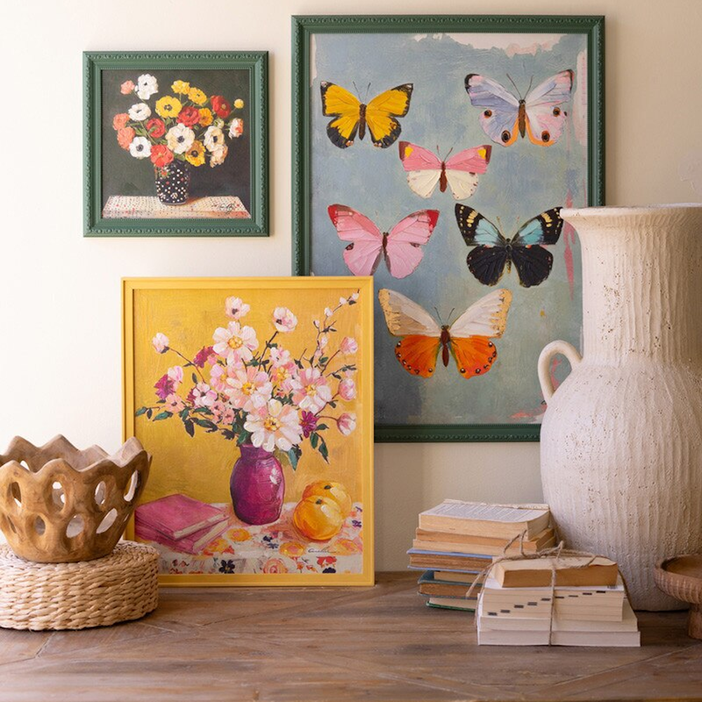 Still life art display with floral and butterfly paintings in yellow and green frames on a rustic wooden table.