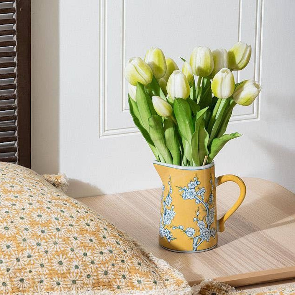 Yellow ceramic jug with blue floral pattern holding a bouquet of white tulips on a wooden table.
