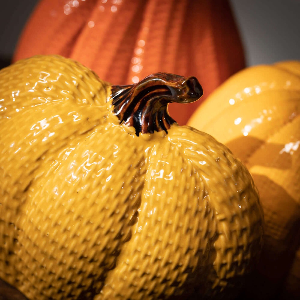 Close-up of a glossy, yellow textured ceramic pumpkin with a dark brown stem, alongside ceramic pumpkins.