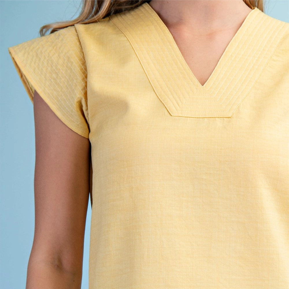 Close-up of a woman in a pale yellow textured linen top featuring detailed pintucks at the V-neck and cap sleeves.