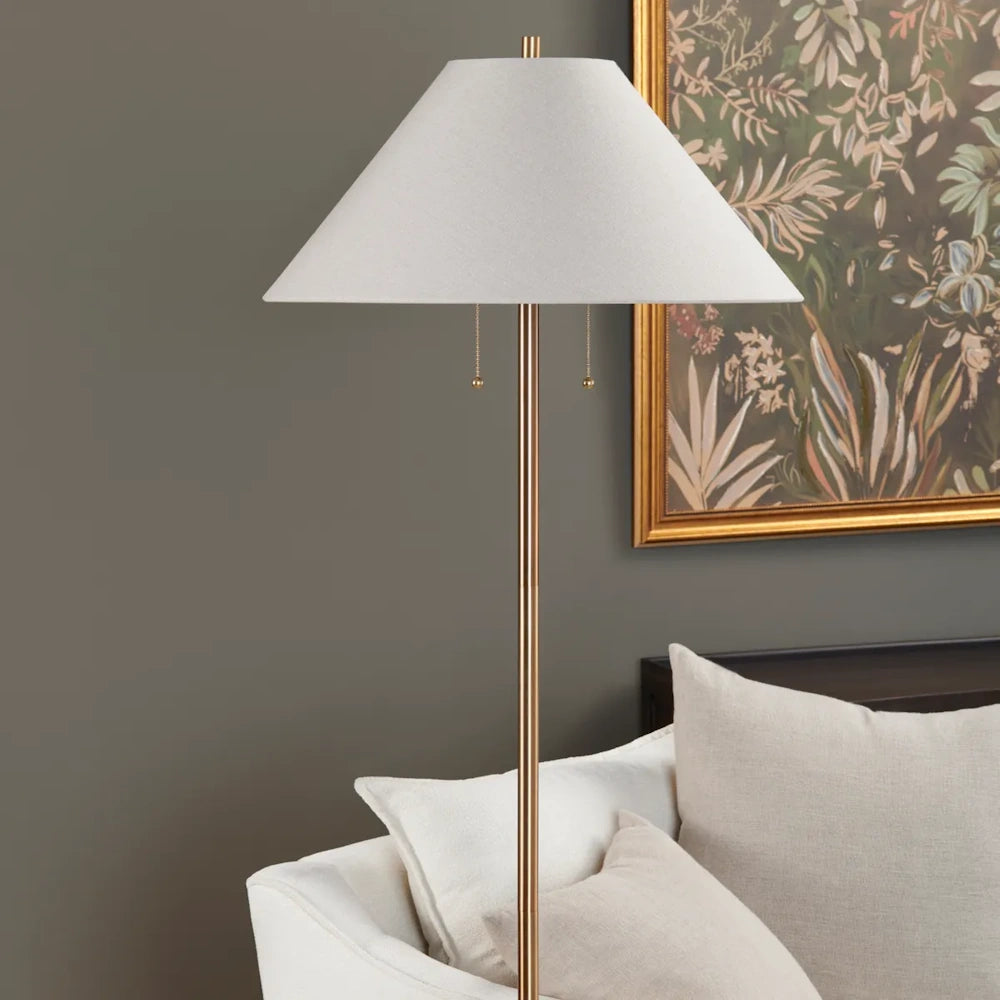Close-up view of a brass floor lamp with a white tapered shade next to a couch in a styled room setting.